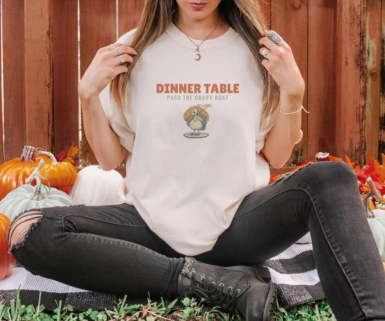 Ivory Comfort Colors t-shirt printed with the words Dinner Table Pass The Gravy Boat and a drawing of a golden roast turkey on a platter, worn by a woman