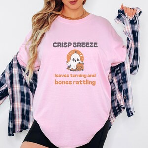 Blossom Comfort Colors t-shirt printed with the words Crisp Breeze Leaves Turning And Bones Rattling and a drawing of a large cute smiling ghost beside a carved jack-o-lantern, worn by a woman