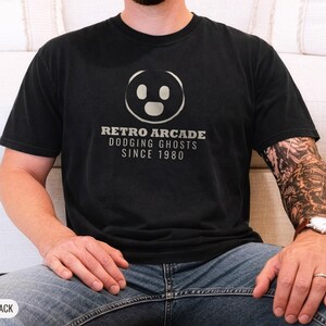 Black Comfort Colors t-shirt printed with the words Retro Arcade Dodging Ghosts Since 1980 and a drawing of a maze with a Pac-Man character, worn by a man