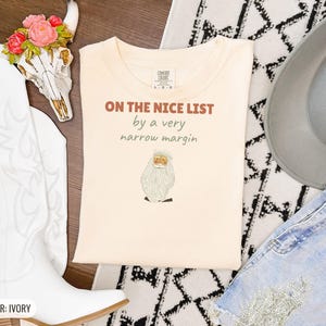 Ivory Comfort Colors t-shirt printed with the words On The Nice List By A Very Narrow Margin and a drawing of a red santa hat beside a candy cane, folded flat lay