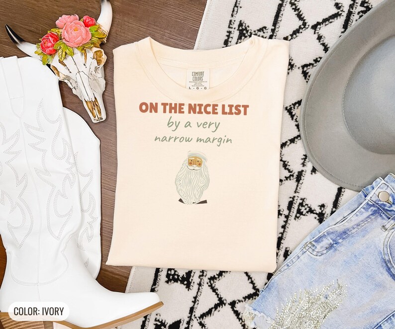 Ivory Comfort Colors t-shirt printed with the words On The Nice List By A Very Narrow Margin and a drawing of a red santa hat beside a candy cane, folded flat lay