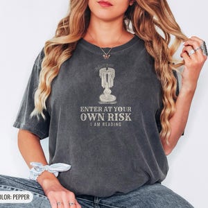 Pepper Comfort Colors t-shirt printed with the words Enter At Your Own Risk I Am Reading and a drawing of a brass candlestick with a single burning candle dripping wax, worn by a woman