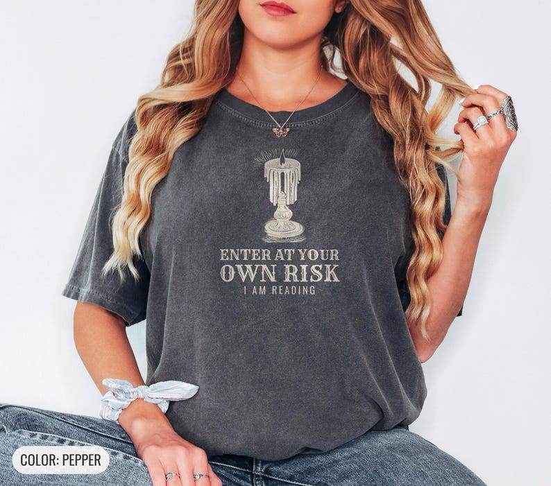 Pepper Comfort Colors t-shirt printed with the words Enter At Your Own Risk I Am Reading and a drawing of a brass candlestick with a single burning candle dripping wax, worn by a woman
