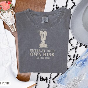 Pepper Comfort Colors t-shirt printed with the words Enter At Your Own Risk I Am Reading and a drawing of a brass candlestick with a single burning candle dripping wax, folded flat lay