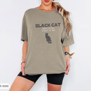 Khaki Comfort Colors t-shirt printed with the words Black Cat Sweet To Me, Spooky To You and a drawing of a fluffy cat that is SOLID JET BLACK from nose to tail, whole body and chest black, sittin, worn by a woman