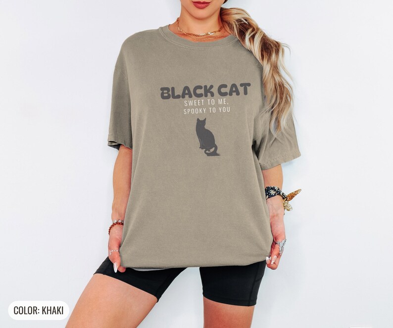 Khaki Comfort Colors t-shirt printed with the words Black Cat Sweet To Me, Spooky To You and a drawing of a fluffy cat that is SOLID JET BLACK from nose to tail, whole body and chest black, sittin, worn by a woman