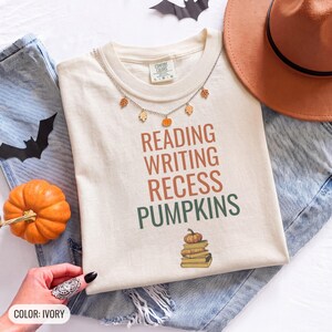 Ivory Comfort Colors t-shirt printed with the words Reading Writing Recess Pumpkins and a drawing of a stack of three vintage schoolbooks with a small orange pumpkin sitting on top, worn by a woman
