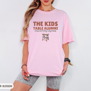 Blossom Comfort Colors t-shirt printed with the words The Kids Table Alumni Still Sitting Together and a drawing of a low wooden folding stool, worn by a woman