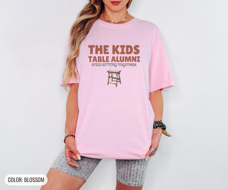 Blossom Comfort Colors t-shirt printed with the words The Kids Table Alumni Still Sitting Together and a drawing of a low wooden folding stool, worn by a woman