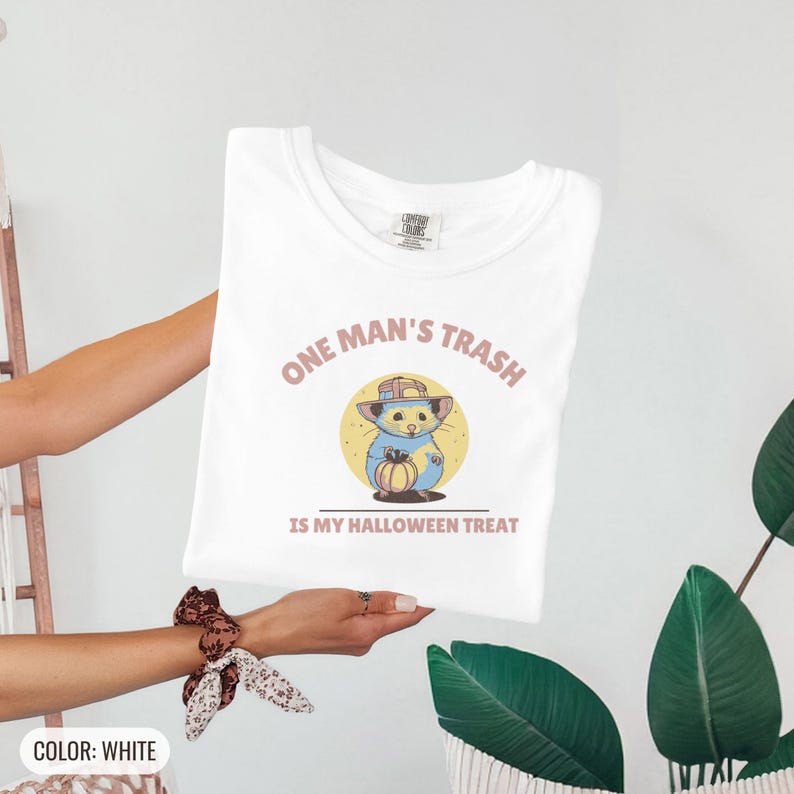 White Comfort Colors t-shirt printed with the words One Man'S Trash Is My Halloween Treat and a drawing of a scruffy opossum wearing a tiny plastic orange pumpkin trick-or-treat bucket on its head, folded flat lay