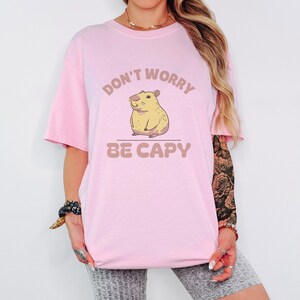 Blossom Comfort Colors t-shirt printed with the words Don'T Worry Be Capy and a drawing of a happy capybara with a small orange citrus slice on its head beside a green lily pad, worn by a woman