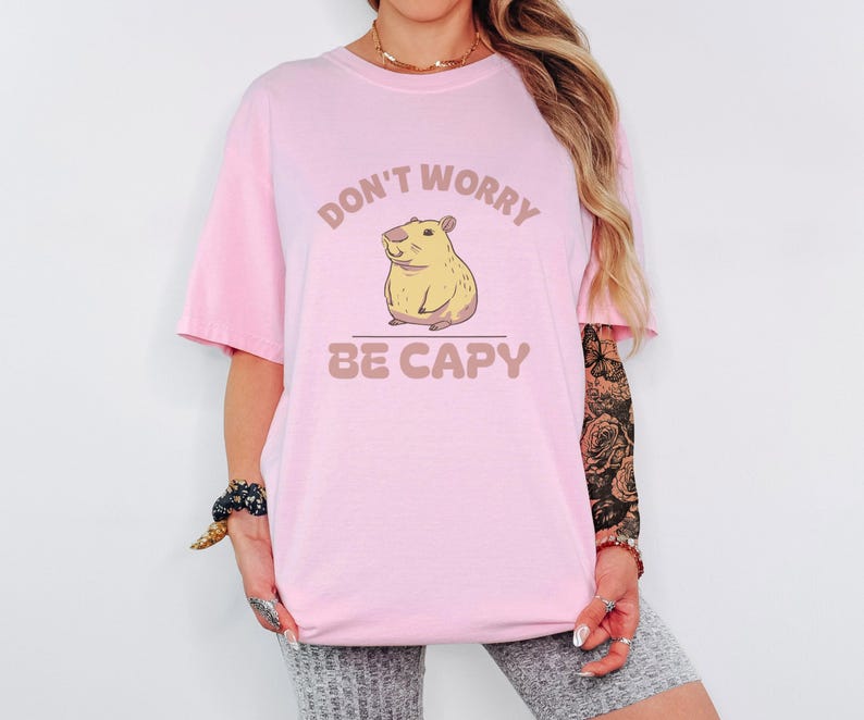 Blossom Comfort Colors t-shirt printed with the words Don'T Worry Be Capy and a drawing of a happy capybara with a small orange citrus slice on its head beside a green lily pad, worn by a woman