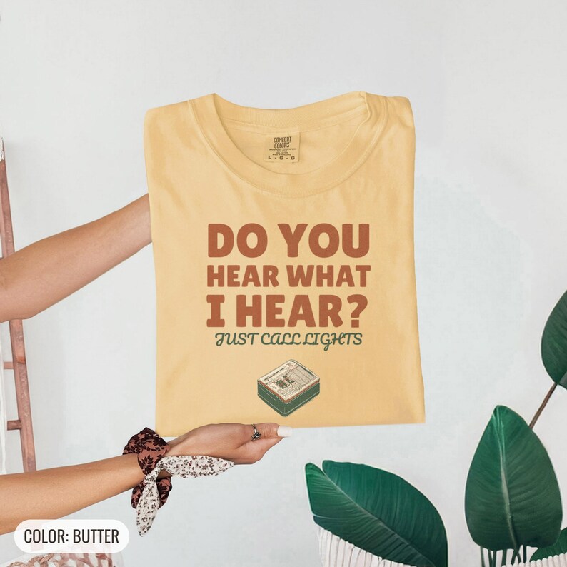 Butter Comfort Colors t-shirt printed with the words Do You Hear What I Hear? Just Call Lights and a drawing of a rectangular electronic medical pager with a small holly sprig beside it, folded flat lay