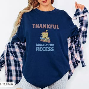True Navy Comfort Colors t-shirt printed with the words Thankful Mostly For Recess and a drawing of a stack of retro school books topped with a glossy red apple and a steaming ceramic coffee, worn by a woman