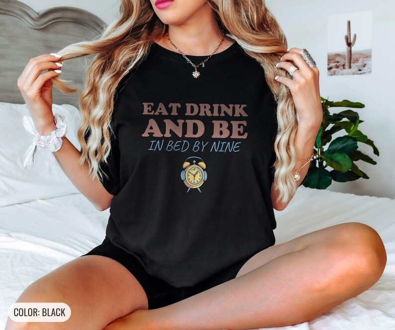 Black Comfort Colors t-shirt printed with the words Eat Drink And Be In Bed By Nine and a drawing of a retro brass wind-up alarm clock with its bells wrapped in fuzzy winter earmuffs, worn by a woman