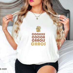 Ivory Comfort Colors t-shirt printed with the words Cookies Cocoa Carols Chaos and a drawing of a retro metal hand mixer with steel beaters, worn by a woman