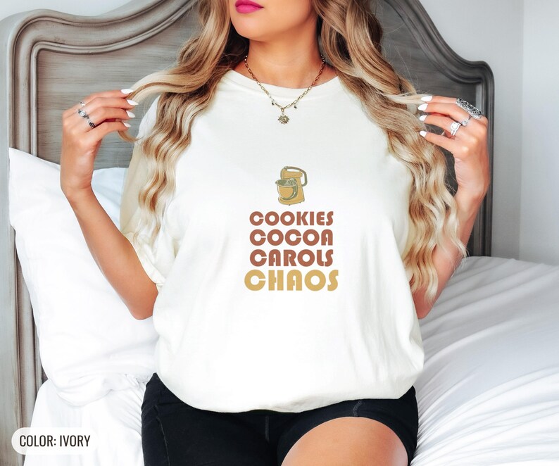 Ivory Comfort Colors t-shirt printed with the words Cookies Cocoa Carols Chaos and a drawing of a retro metal hand mixer with steel beaters, worn by a woman