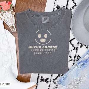 Pepper Comfort Colors t-shirt printed with the words Retro Arcade Dodging Ghosts Since 1980 and a drawing of a maze with a Pac-Man character, folded flat lay