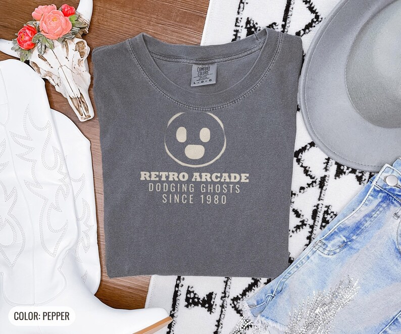 Pepper Comfort Colors t-shirt printed with the words Retro Arcade Dodging Ghosts Since 1980 and a drawing of a maze with a Pac-Man character, folded flat lay