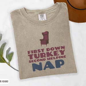 Khaki Comfort Colors t-shirt printed with the words First Down Turkey Second Helping Nap and a drawing of a vintage brown recliner with a footrest extended outward, folded flat lay