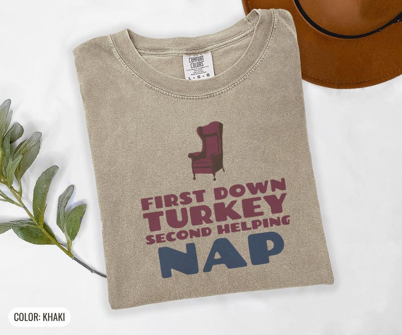 Khaki Comfort Colors t-shirt printed with the words First Down Turkey Second Helping Nap and a drawing of a vintage brown recliner with a footrest extended outward, folded flat lay