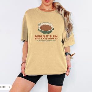 Butter Comfort Colors t-shirt printed with the words What'S In The Casserole? Don'T Ask Questions and a drawing of a bubbly baked casserole in a rectangular ceramic baking dish topped with crispy fried oni, worn by a woman