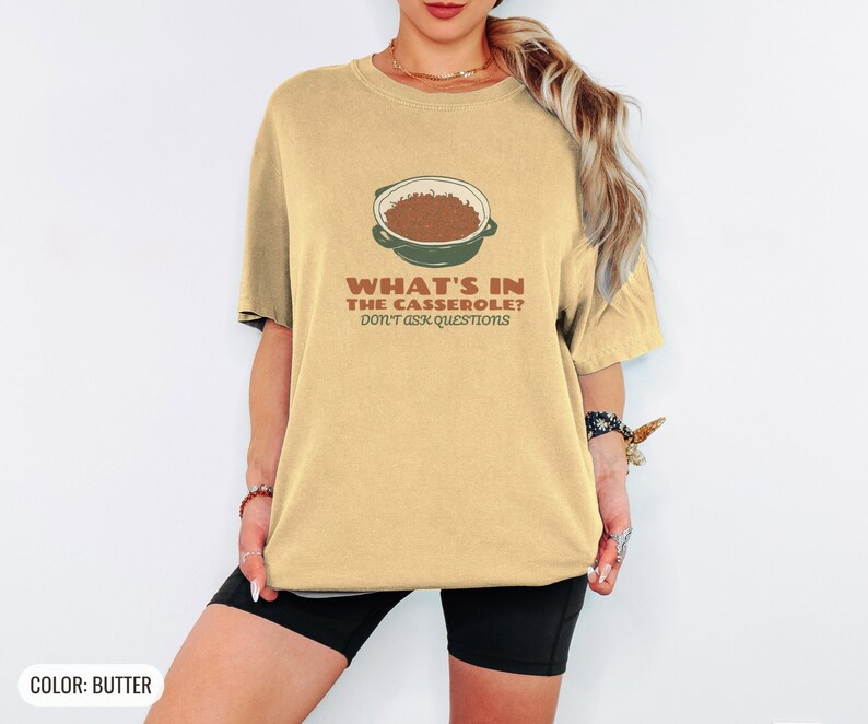 Butter Comfort Colors t-shirt printed with the words What'S In The Casserole? Don'T Ask Questions and a drawing of a bubbly baked casserole in a rectangular ceramic baking dish topped with crispy fried oni, worn by a woman