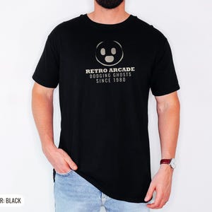 Black Comfort Colors t-shirt printed with the words Retro Arcade Dodging Ghosts Since 1980 and a drawing of a maze with a Pac-Man character, worn by a man