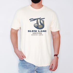 Ivory Comfort Colors t-shirt printed with the words Slow Lane Arriving Whenever I Arrive and a drawing of a sloth hanging upside down from a branch with a peaceful smile, worn by a man