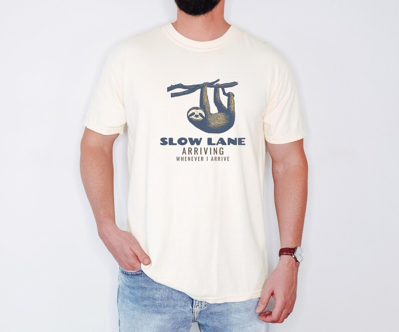 Ivory Comfort Colors t-shirt printed with the words Slow Lane Arriving Whenever I Arrive and a drawing of a sloth hanging upside down from a branch with a peaceful smile, worn by a man