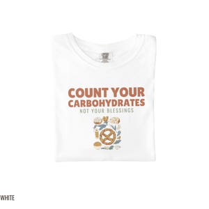 White Comfort Colors t-shirt printed with the words Count Your Carbohydrates Not Your Blessings and a drawing of a golden-brown braided pretzel with coarse salt grains, folded flat lay