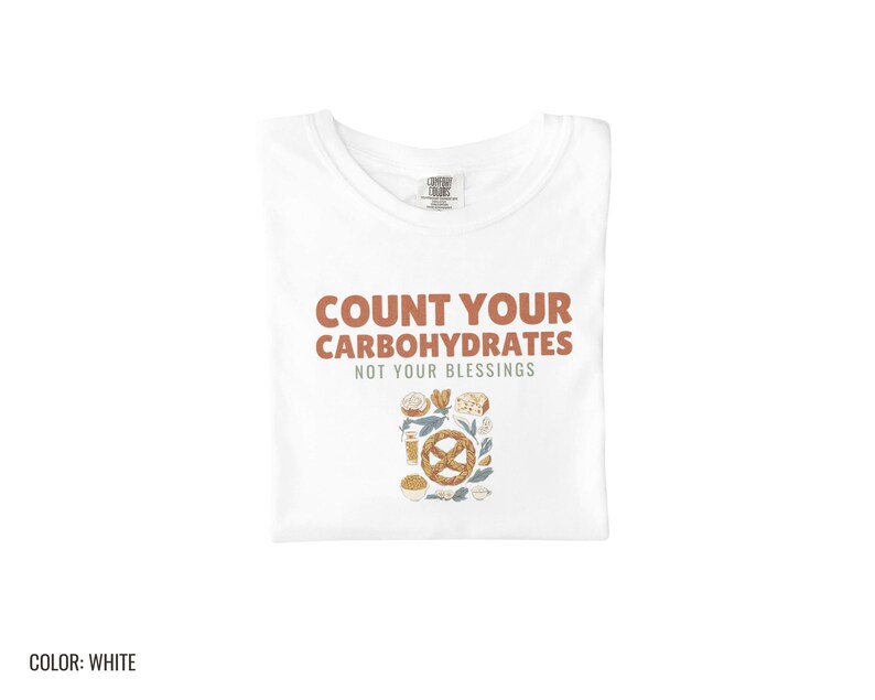 White Comfort Colors t-shirt printed with the words Count Your Carbohydrates Not Your Blessings and a drawing of a golden-brown braided pretzel with coarse salt grains, folded flat lay