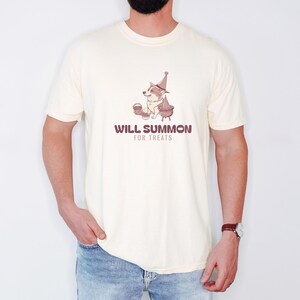 Ivory Comfort Colors t-shirt printed with the words Will Summon For Treats and a drawing of a cute corgi wearing a wizard hat sitting next to a bubbling cauldron overflowing with bon, worn by a man