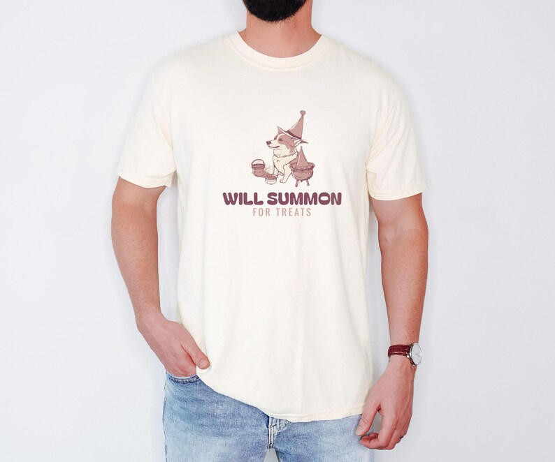 Ivory Comfort Colors t-shirt printed with the words Will Summon For Treats and a drawing of a cute corgi wearing a wizard hat sitting next to a bubbling cauldron overflowing with bon, worn by a man