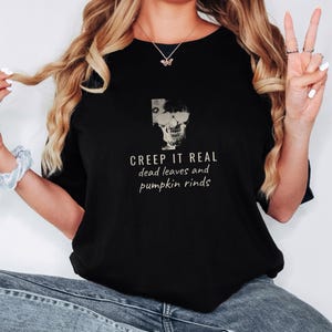 Black Comfort Colors t-shirt printed with the words Creep It Real Dead Leaves And Pumpkin Rinds and a drawing of a grinning skull with black sunglasses, worn by a woman
