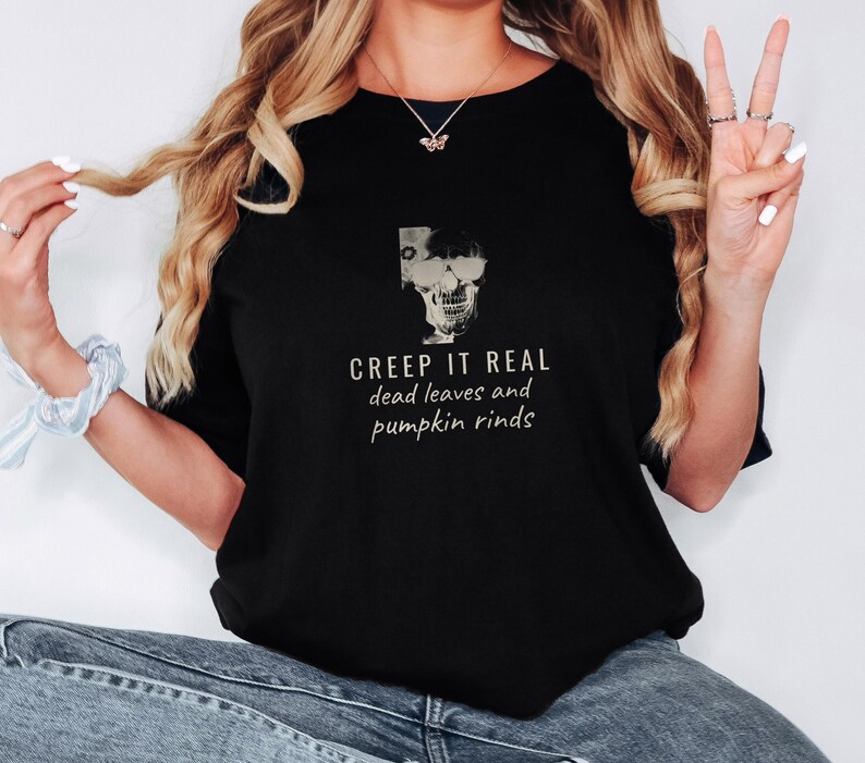 Black Comfort Colors t-shirt printed with the words Creep It Real Dead Leaves And Pumpkin Rinds and a drawing of a grinning skull with black sunglasses, worn by a woman
