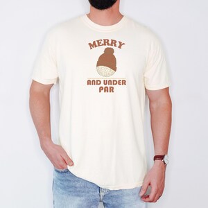 Ivory Comfort Colors t-shirt printed with the words Merry And Under Par and a drawing of a dimpled golf ball wearing a tiny red and white striped knit winter beanie with a green p, worn by a man