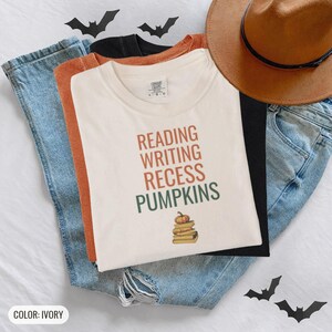 Ivory Comfort Colors t-shirt printed with the words Reading Writing Recess Pumpkins and a drawing of a stack of three vintage schoolbooks with a small orange pumpkin sitting on top, worn by a woman
