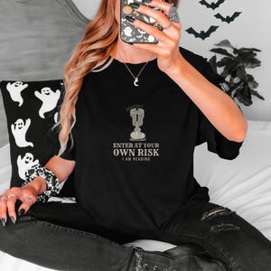 Black Comfort Colors t-shirt printed with the words Enter At Your Own Risk I Am Reading and a drawing of a brass candlestick with a single burning candle dripping wax, worn by a woman