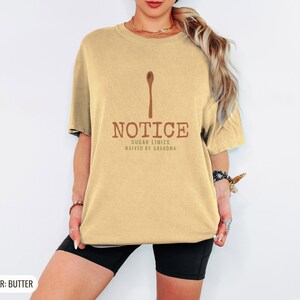 Butter Comfort Colors t-shirt printed with the words Notice Sugar Limits Waived By Grandma and a drawing of a painted wooden spoon crossed over a cinnamon stick bundle, worn by a woman