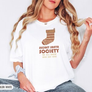 White Comfort Colors t-shirt printed with the words Secret Santa Society We All Know Who Got Who and a drawing of a knitted wool stocking stuffed with stick candy, worn by a woman