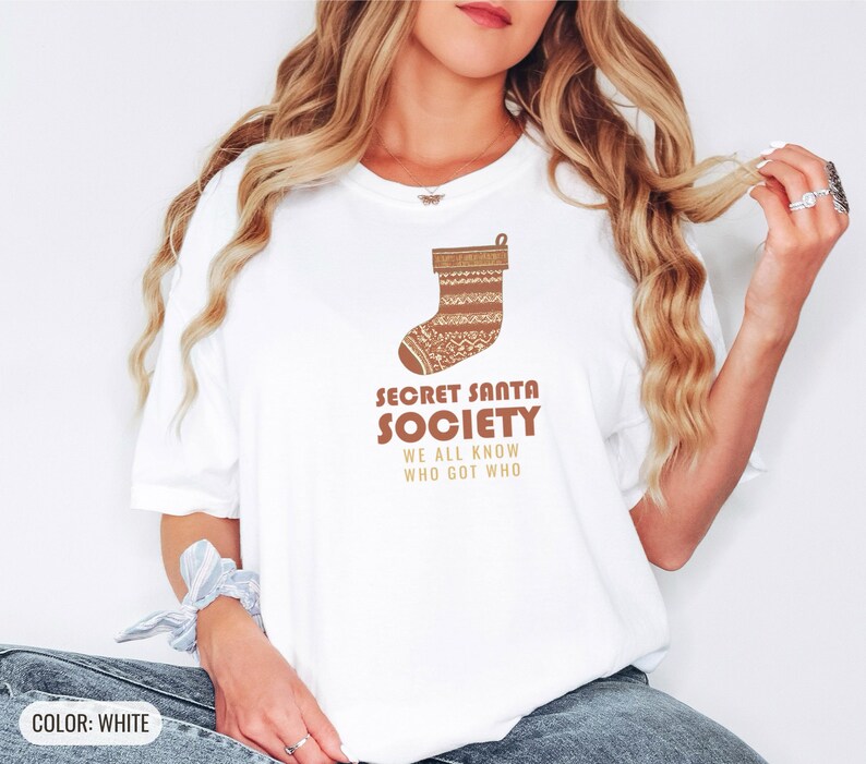 White Comfort Colors t-shirt printed with the words Secret Santa Society We All Know Who Got Who and a drawing of a knitted wool stocking stuffed with stick candy, worn by a woman