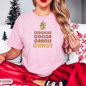 Blossom Comfort Colors t-shirt printed with the words Cookies Cocoa Carols Chaos and a drawing of a retro metal hand mixer with steel beaters, worn by a woman