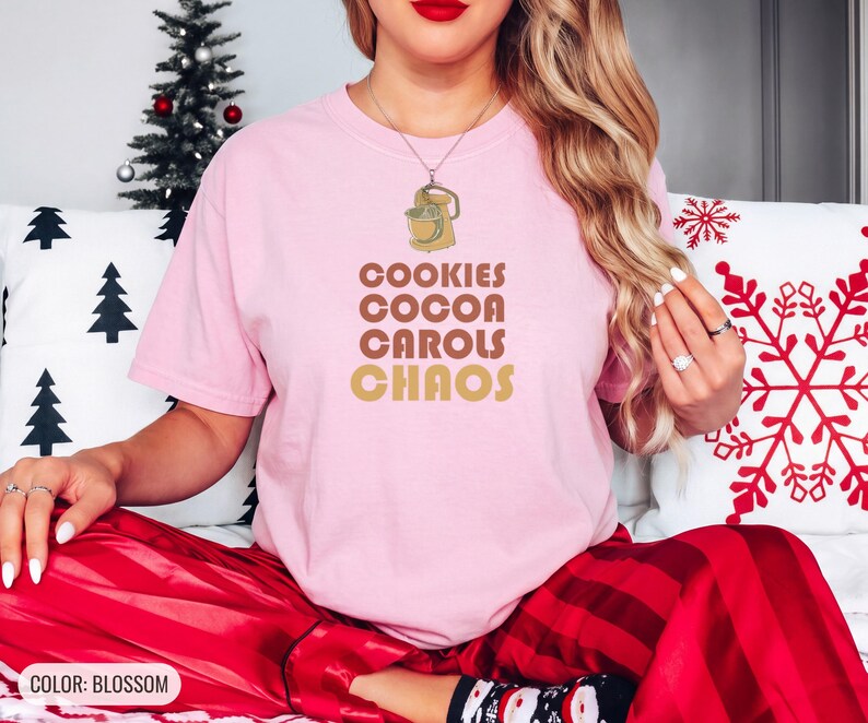 Blossom Comfort Colors t-shirt printed with the words Cookies Cocoa Carols Chaos and a drawing of a retro metal hand mixer with steel beaters, worn by a woman