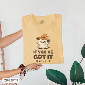 Butter Comfort Colors t-shirt printed with the words If You'Ve Got It Haunt It and a drawing of a cute retro cartoon ghost wearing a witch hat, folded flat lay