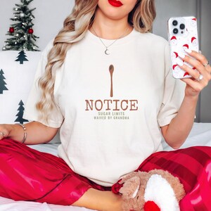 Ivory Comfort Colors t-shirt printed with the words Notice Sugar Limits Waived By Grandma and a drawing of a painted wooden spoon crossed over a cinnamon stick bundle, worn by a woman