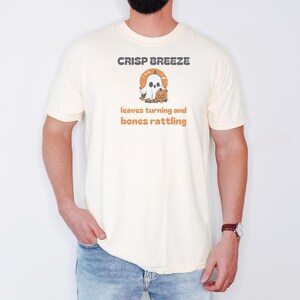 Ivory Comfort Colors t-shirt printed with the words Crisp Breeze Leaves Turning And Bones Rattling and a drawing of a large cute smiling ghost beside a carved jack-o-lantern, worn by a man
