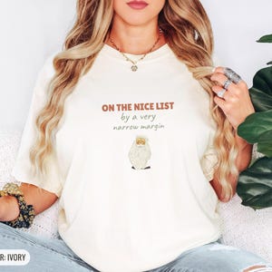 Ivory Comfort Colors t-shirt printed with the words On The Nice List By A Very Narrow Margin and a drawing of a red santa hat beside a candy cane, worn by a woman