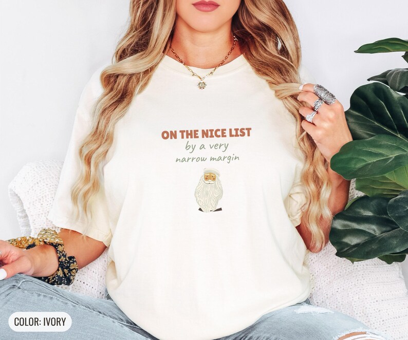 Ivory Comfort Colors t-shirt printed with the words On The Nice List By A Very Narrow Margin and a drawing of a red santa hat beside a candy cane, worn by a woman