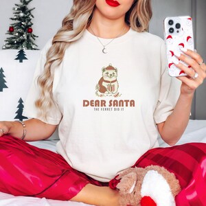 Ivory Comfort Colors t-shirt printed with the words Dear Santa The Ferret Did It and a drawing of a mischievous ferret dragging a striped Christmas stocking filled with shiny ornaments, worn by a woman